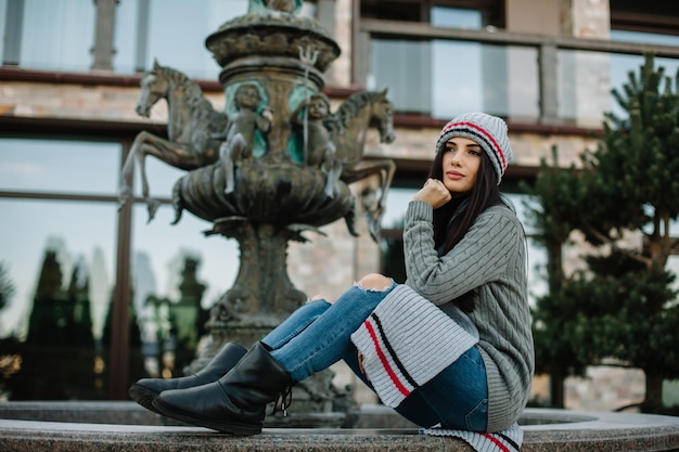 Una niña vestida con una gorra y una bufanda se sienta junto a una fuente al aire libre