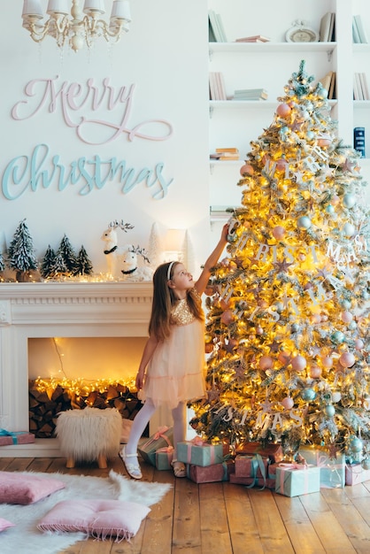 niña, vestida con un hermoso vestido blanco de moda, posa cerca del árbol de Navidad.