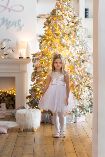 niña, vestida con un hermoso vestido blanco de moda, posa cerca del árbol de Navidad.