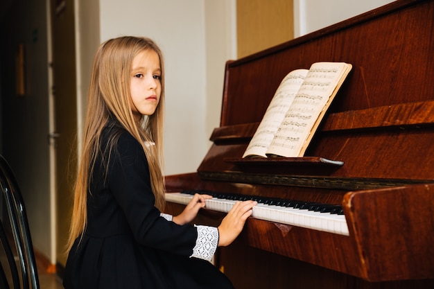 Niña vestida de negro aprende a tocar el piano. El niño toca un instrumento musical. Colegiala mirando hacia un lado.