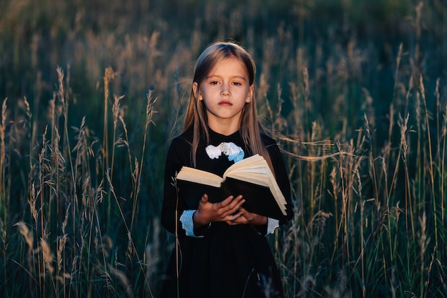 Una niña vestida de negro se para en la hierba alta y sostiene un libro verde a la luz del sol poniente. Un niño con una expresión facial pensativa.