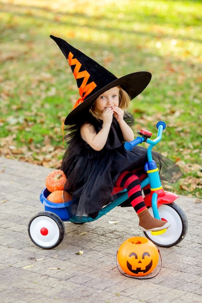 Niña vestida con traje de bruja en bicicleta con calabazas y dulces