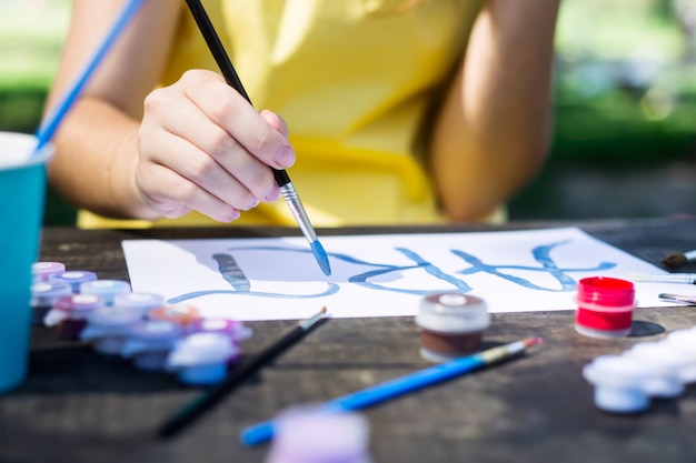 Una niña en un vestido amarillo escribe pinturas en una hoja de papel la palabra arte. En una mesa de madera. Concepto de hobby creativo