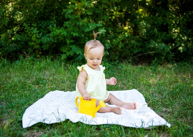Una niña con un vestido amarillo se sienta sobre una manta blanca con una regadera en la naturaleza