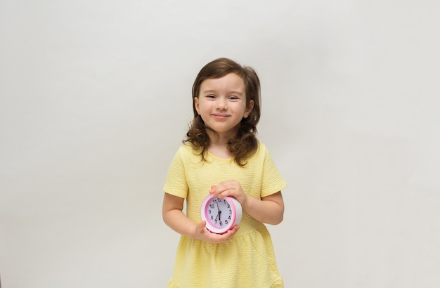 Una niña con un vestido amarillo sostiene un reloj despertador de mesa sobre un fondo blanco con un lugar para el texto