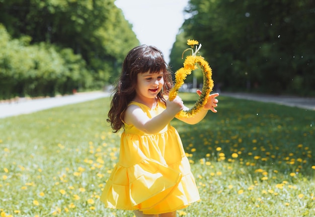 Una niña con un vestido amarillo de verano atrapa burbujas de jabón en la hierba en la corona de dientes de león del parque