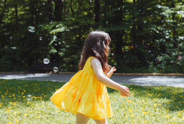 Una niña con un vestido amarillo de verano atrapa burbujas de jabón en la hierba en la corona de dientes de león del parque