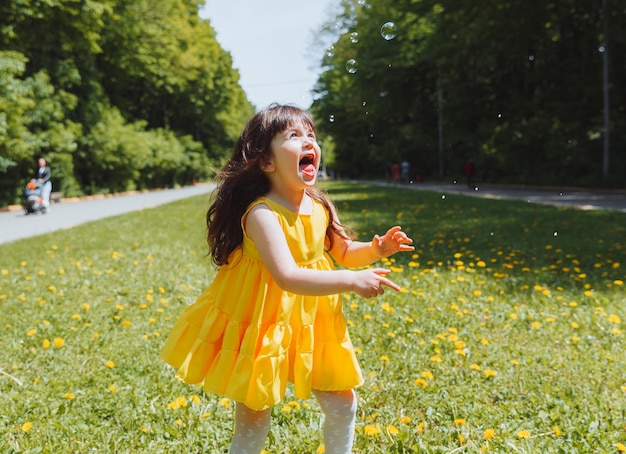 Una niña con un vestido amarillo de verano atrapa burbujas de jabón en la hierba en la corona de dientes de león del parque