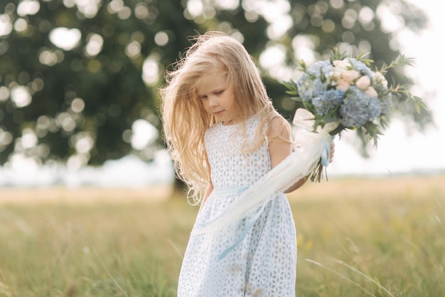 Niña en vestido azul cielo con soporte de ramo en el campo frente a un gran árbol Niño sonríe y diviértete