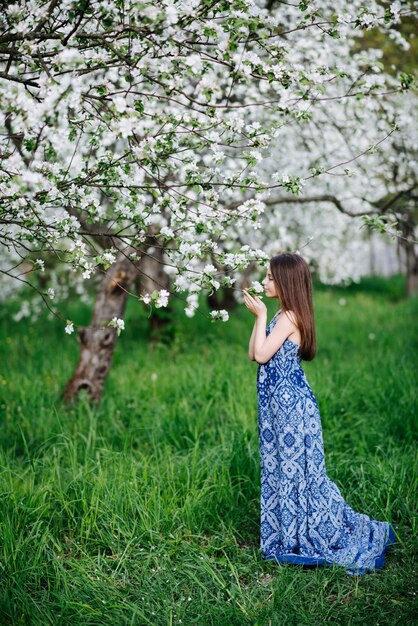 Una niña con un vestido azul largo disfruta del aroma de los manzanos en flor en el jardín. Jardín floreciente. Temporada de alergias.