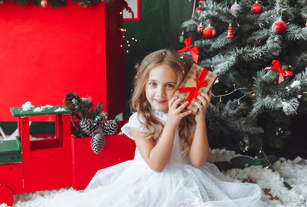 Una niña con un vestido blanco está sentada en el suelo y sosteniendo un regalo de Año Nuevo en el fondo de un árbol de Navidad el concepto de Navidad y año nuevo