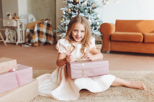 Una niña con un vestido blanco está sentada en el suelo y sosteniendo un regalo de Año Nuevo en el fondo de un árbol de Navidad el concepto de Navidad y año nuevo