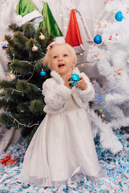 Niña con vestido blanco juega cerca del árbol de Navidad