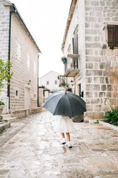 Niña con un vestido blanco de pie afuera bajo un paraguas negro durante la lluvia
