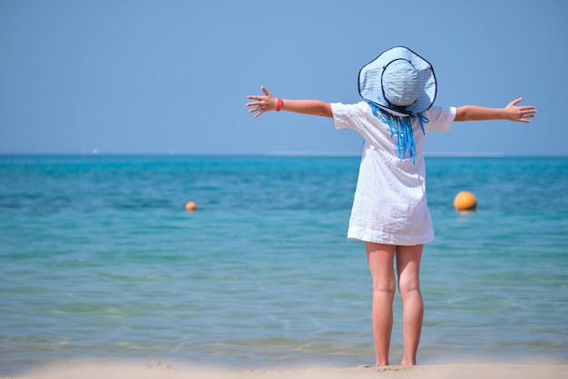 Niña con vestido blanco y sombrero descalzo con las manos extendidas en la playa disfrutando de vacaciones tropicales mirando el agua del océano