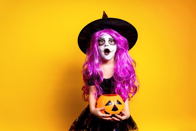 Niña en vestido de bruja de Halloween y sombrero asustando y haciendo muecas aisladas sobre fondo amarillo. Truco o trato.