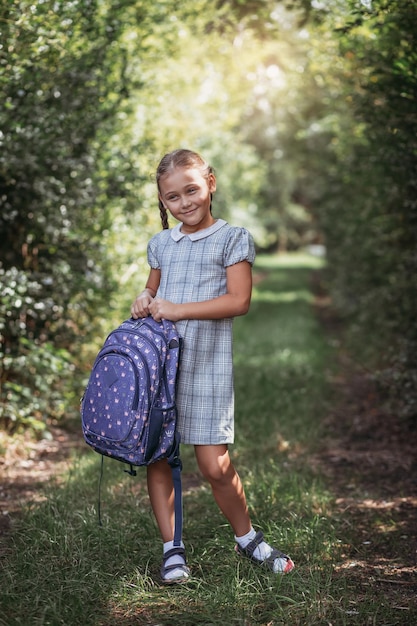 Una niña con un vestido con coletas sostiene una gran mochila azul y camina por el callejón de la carretera El primer día de septiembre en la escuela primaria