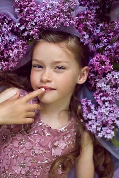 Niña con un vestido de flores púrpura yace en el suelo entre lilas sobre un velo en primavera