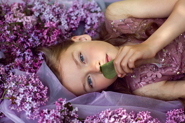 Niña con un vestido de flores púrpura yace en el suelo entre lilas sobre un velo en primavera