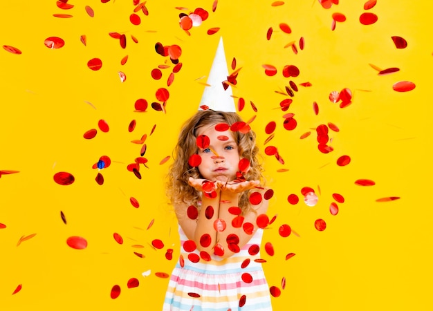 Una niña con un vestido y una gorra festiva se encuentra sobre un fondo amarillo con confeti y sonrisas