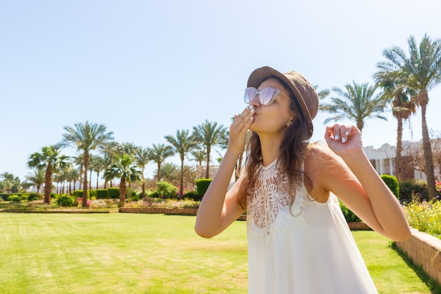 Una niña con un vestido largo blanco está caminando sobre una palma tropical y envía un beso a la cámara.