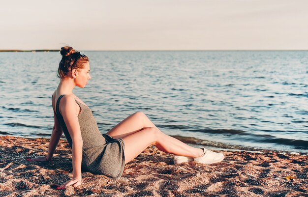 Niña con un vestido ligero y gafas de sol sentado en la arena y mirando al mar