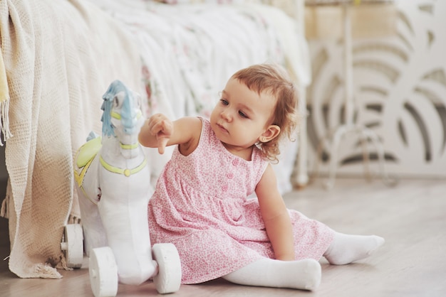 Niña en vestido lindo emplazamiento en la cama jugando con juguetes en el hogar. Habitación infantil blanca vintage. Concepto de infancia