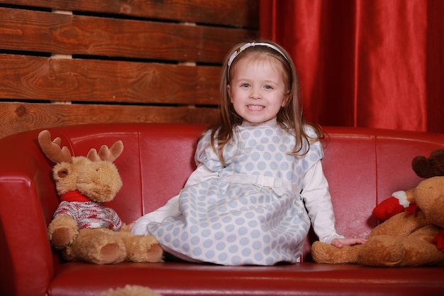 Niña con vestido de lunares blancos divirtiéndose en el sofá rojo en el estudio de Navidad. Árbol de Navidad, oso de peluche y caja de regalos en la parte delantera.