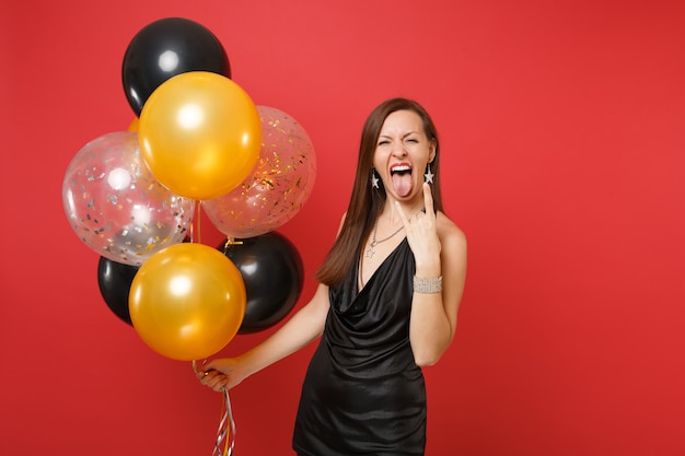 Niña con vestido negro celebrando mostrando la lengua, cartel de rock-n-roll con globos de aire aislados sobre fondo rojo. Concepto de fiesta de vacaciones de maqueta de cumpleaños de feliz año nuevo de San Valentín.