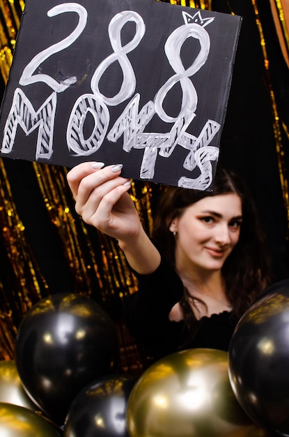 Una niña con un vestido negro sobre un fondo dorado sostiene globos en sus manos.