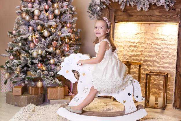 Niña en vestido paseos a caballo de madera cerca del árbol de Navidad en la sala de estar