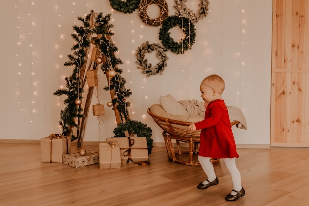 Niña en vestido rojo ejecuta minimalismo ligero estilo apartamento. niño se encuentra en la pared blanca con guirnaldas Árbol de Navidad con regalos envueltos. Navidad Año nuevo interior Celebración del día de San Valentín