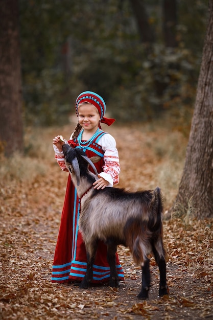 una niña con un vestido rojo se encuentra junto a un cabrito negro en el bosque de otoño