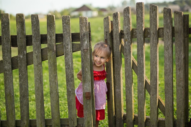 Una niña con un vestido rojo mira desde detrás de una valla de madera.