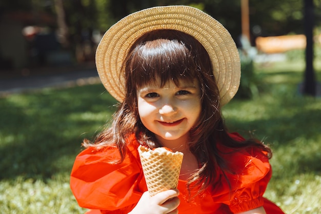 Una niña con un vestido rojo y un sombrero de paja come helado en un cono en el parque