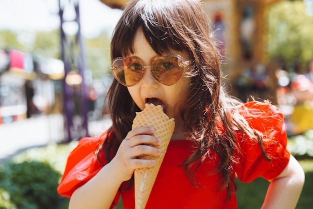 Una niña con un vestido rojo y un sombrero de paja come helado en un cono en el parque