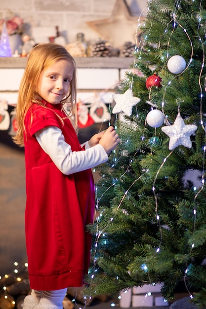 Una niña con un vestido rojo y un sombrero de santa claus cerca de la chimenea se aferra a un juguete en el árbol de navidad