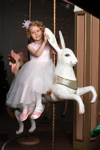 Una niña con un vestido rosa y una corona en un carrusel con una liebre blanca.