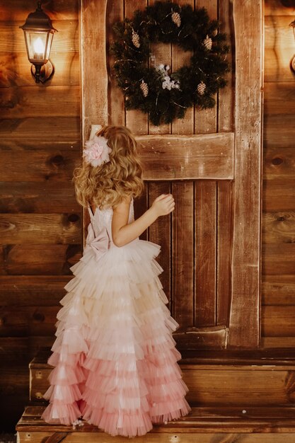 Una niña con un vestido rosa se encuentra frente a una puerta de madera con una corona de Navidad. Bienvenido al cuento de hadas