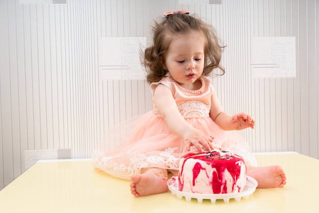 Niña en vestido rosa sentada en la superficie de la mesa y comiendo pastel