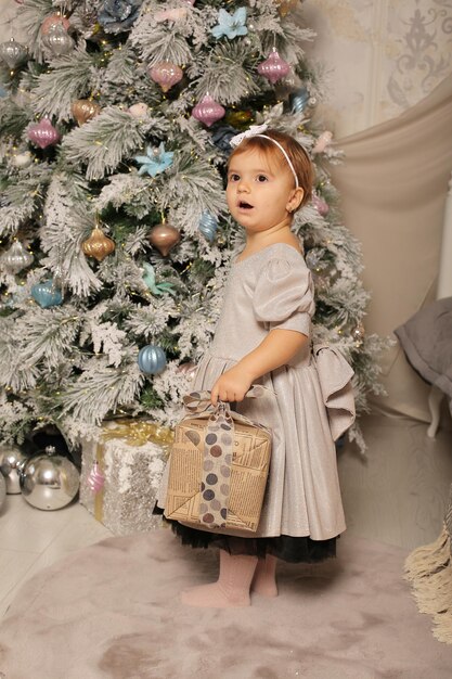 una niña con un vestido sosteniendo una caja de regalo está parada en el árbol de Navidad