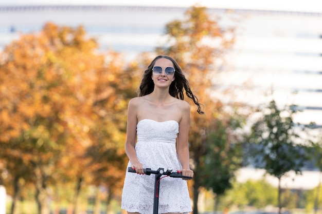 Una niña con un vestido de verano monta un scooter eléctrico en la acera