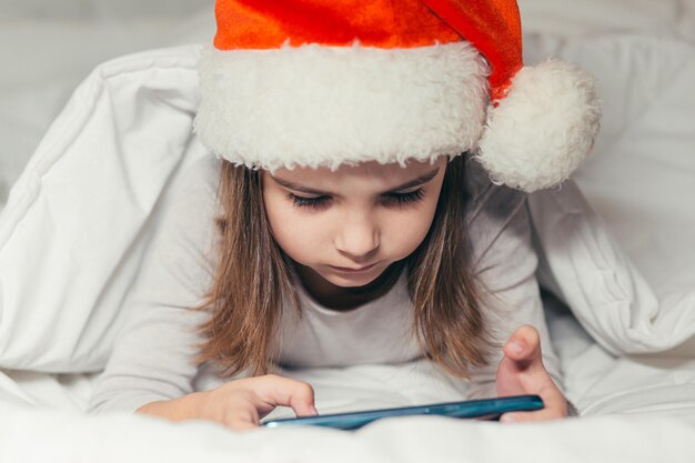 Una niña yace en la cama con un sombrero de Santa Claus y mira dibujos animados, estudia, juega un juego en el teléfono