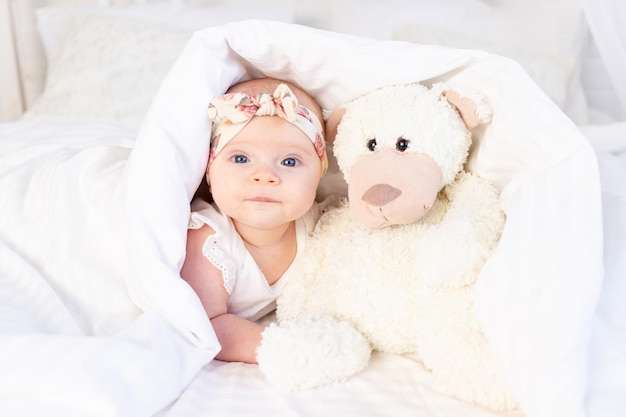 Una niña yace debajo de una manta con un osito de peluche y sonríe en una cama de algodón blanco en casa