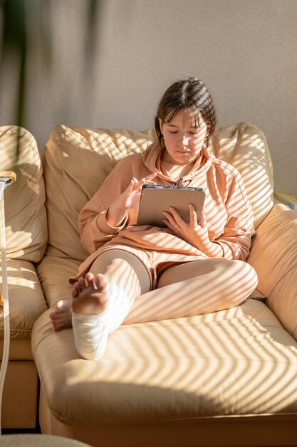 Niña con yeso en la pierna sentada en el sofá y dibujando en una tableta gráfica