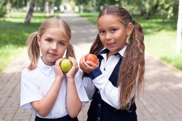 Las niñas de 6 a 7 años sostienen una manzana fresca en sus manos al aire libre. En mochila escolar y uniforme. Infancia. De vuelta a la escuela. 1 de septiembre. Buenos días.