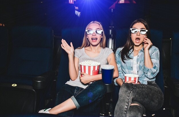 Las niñas asustadas y emocionadas están sentadas en una sala de cine y viendo películas. Están asombrados También las niñas tienen canastas de palomitas de maíz y una gran taza de cola.