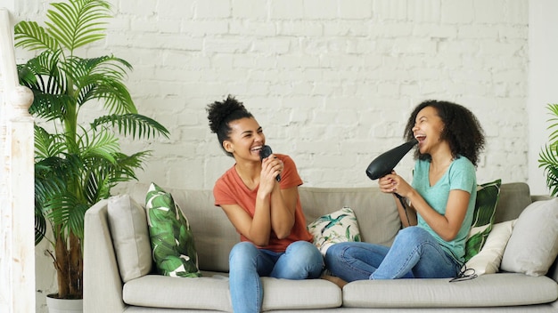 Niñas divertidas de raza mixta bailan cantando con secador de pelo y peine sentado en el sofá