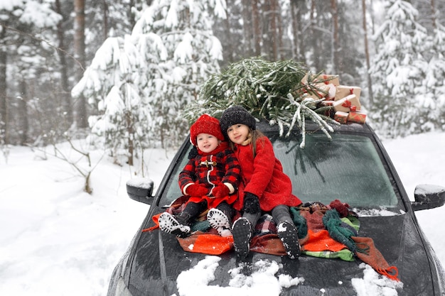 Las niñas en edad preescolar están sentadas en el techo de un automóvil junto a un árbol de Navidad y regalos