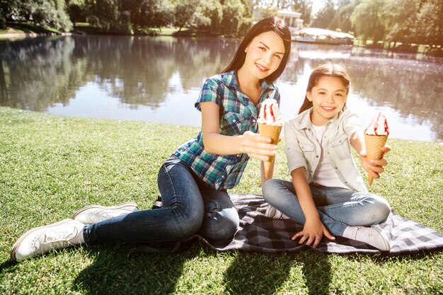 Las niñas están sentadas en una manta cerca del agua y posando. Están sosteniendo contras de helado y mirando a la cámara. Ellos están sonriendo. Hace sol afuera en el parque.